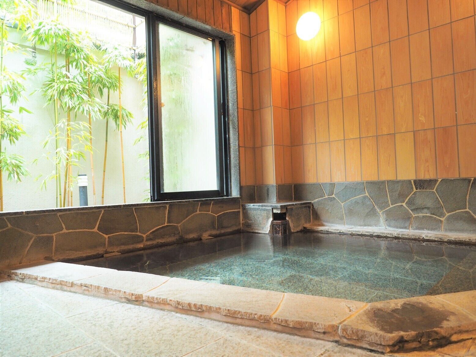 Un baño de aguas termales con paredes de piedra y techo de madera, una ventana con bambú en el exterior y un repisa de piedra.