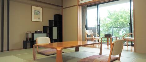 Una habitación con tatami, una mesa baja, dos sillas, una estantería negra y una ventana con vistas a árboles.