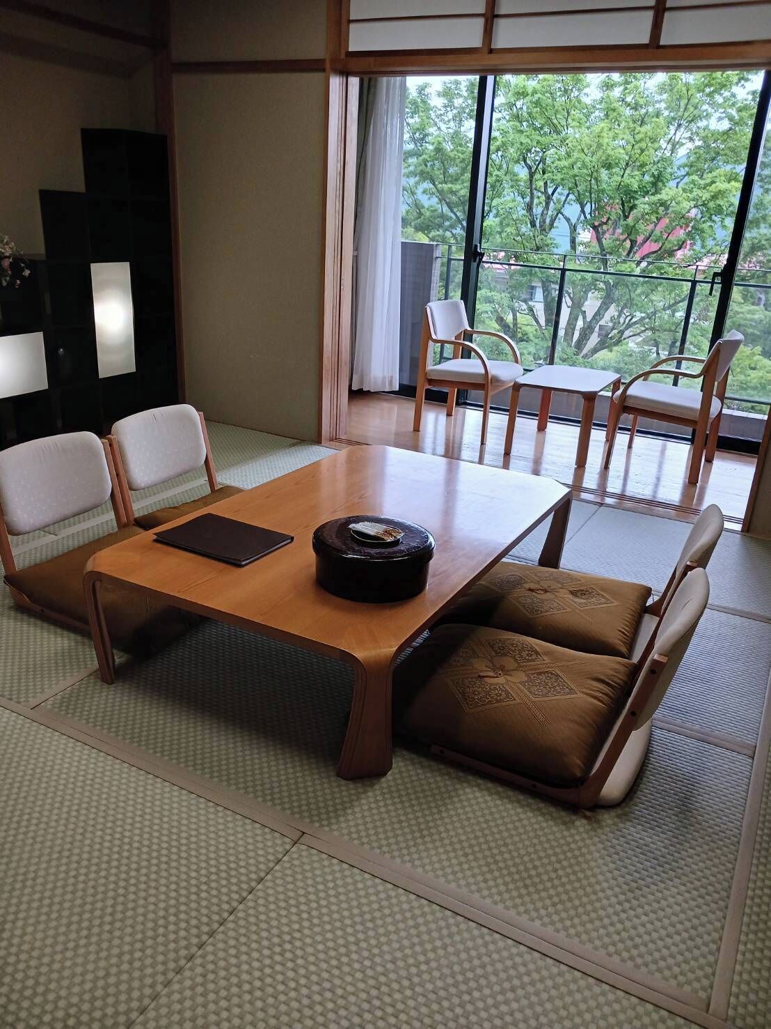 A traditional Japanese-style room with tatami flooring, a low table with cushions, and a view of greenery through large windows.