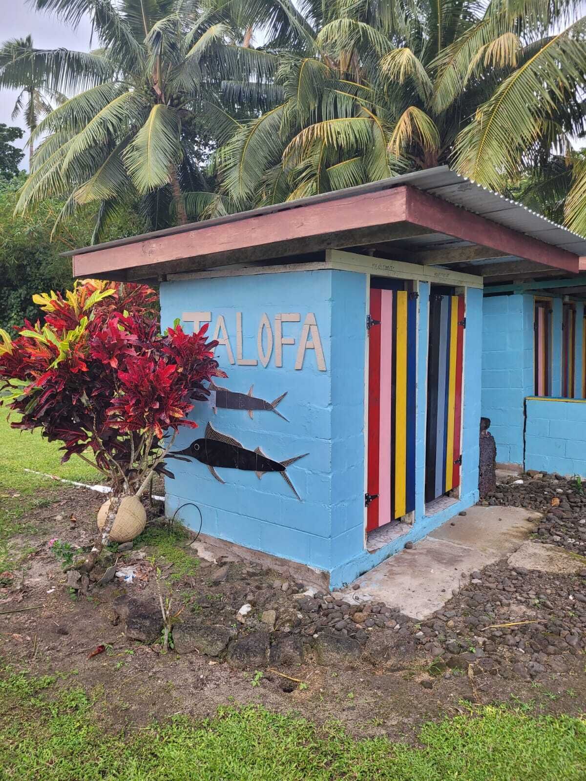 Un petit bâtiment coloré, sur lequel on peut lire le mot « TALOFA », peint sur le côté, et qui est entouré de palmiers.