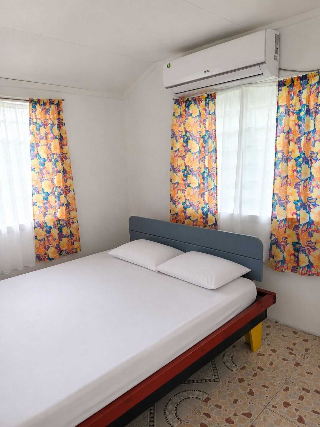Habitación de hotel con cama, unidad de aire acondicionado, cortinas con motivos florales y piso de baldosas.