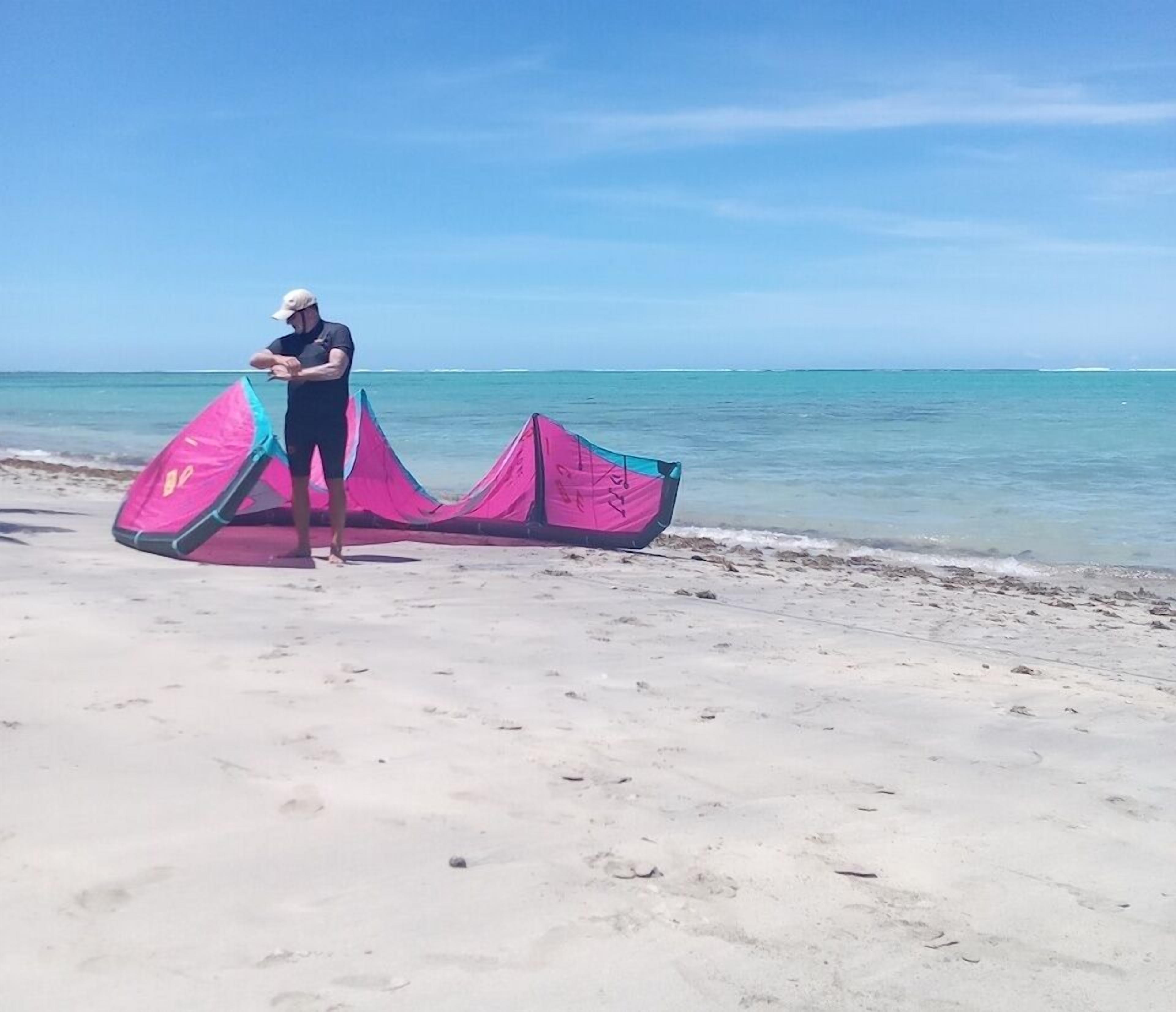 Una playa con una persona de pie junto a una cometa rosa y azul.