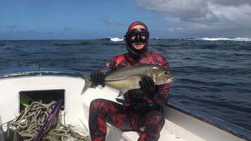 Una persona con traje de neopreno rojo y negro sostiene un pez en un bote con interior blanco y cuerdas en la cubierta.