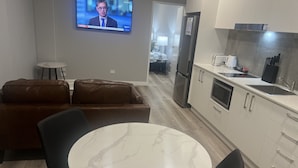 Een moderne keuken met een koelkast, magnetron en gootsteen. Er is een eethoek met een marmeren tafel en stoelen. Een televisie is aan de muur bevestigd en laat een nieuwsuitzending zien.