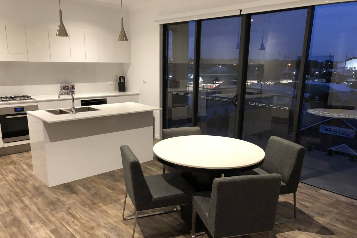 A modern kitchen with a central island, a dining area with a round table and chairs, and large windows offering a view of the cityscape.