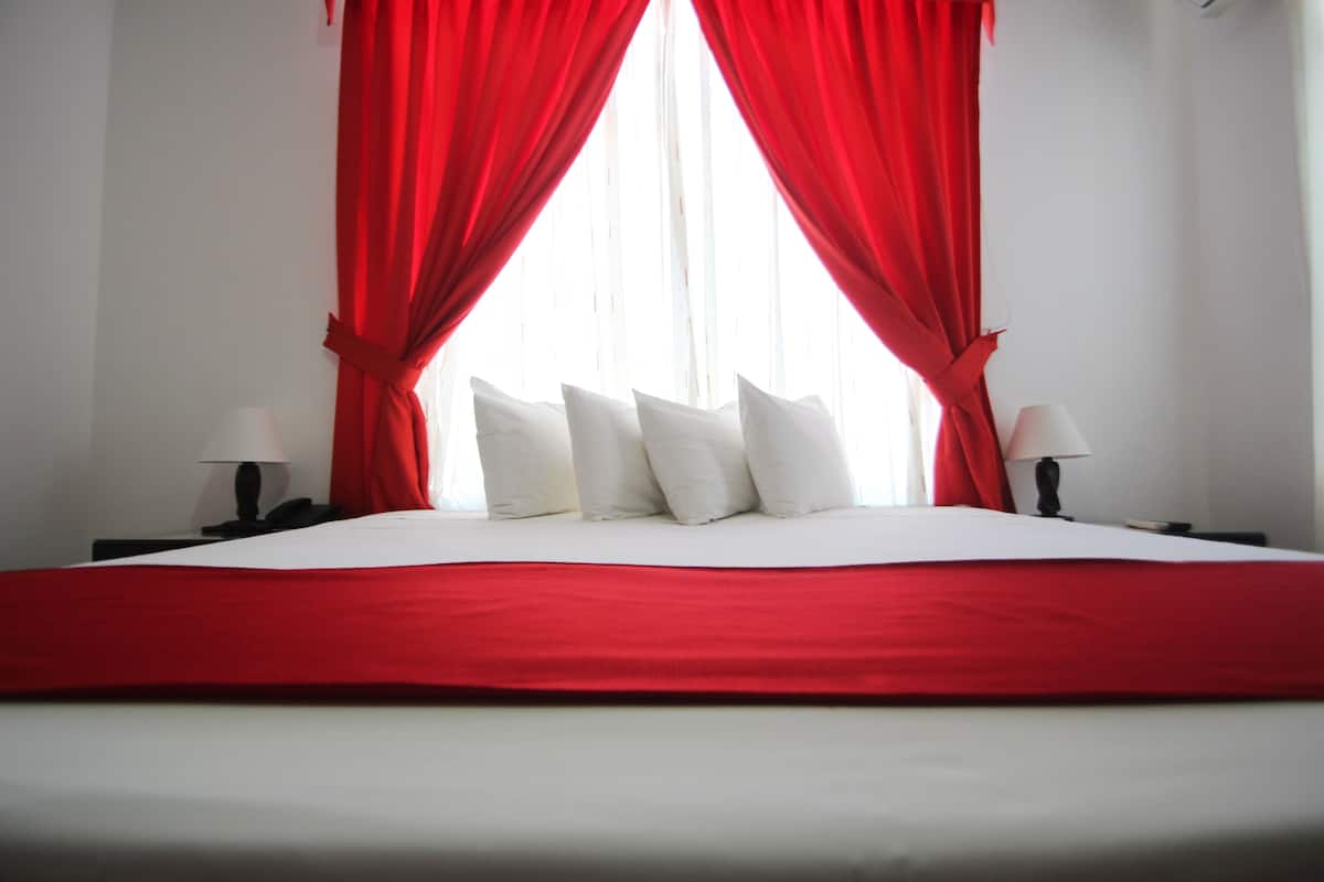 A neatly made bed with white bedding and red curtains, two bedside tables with lamps, and a window with sheer white curtains.