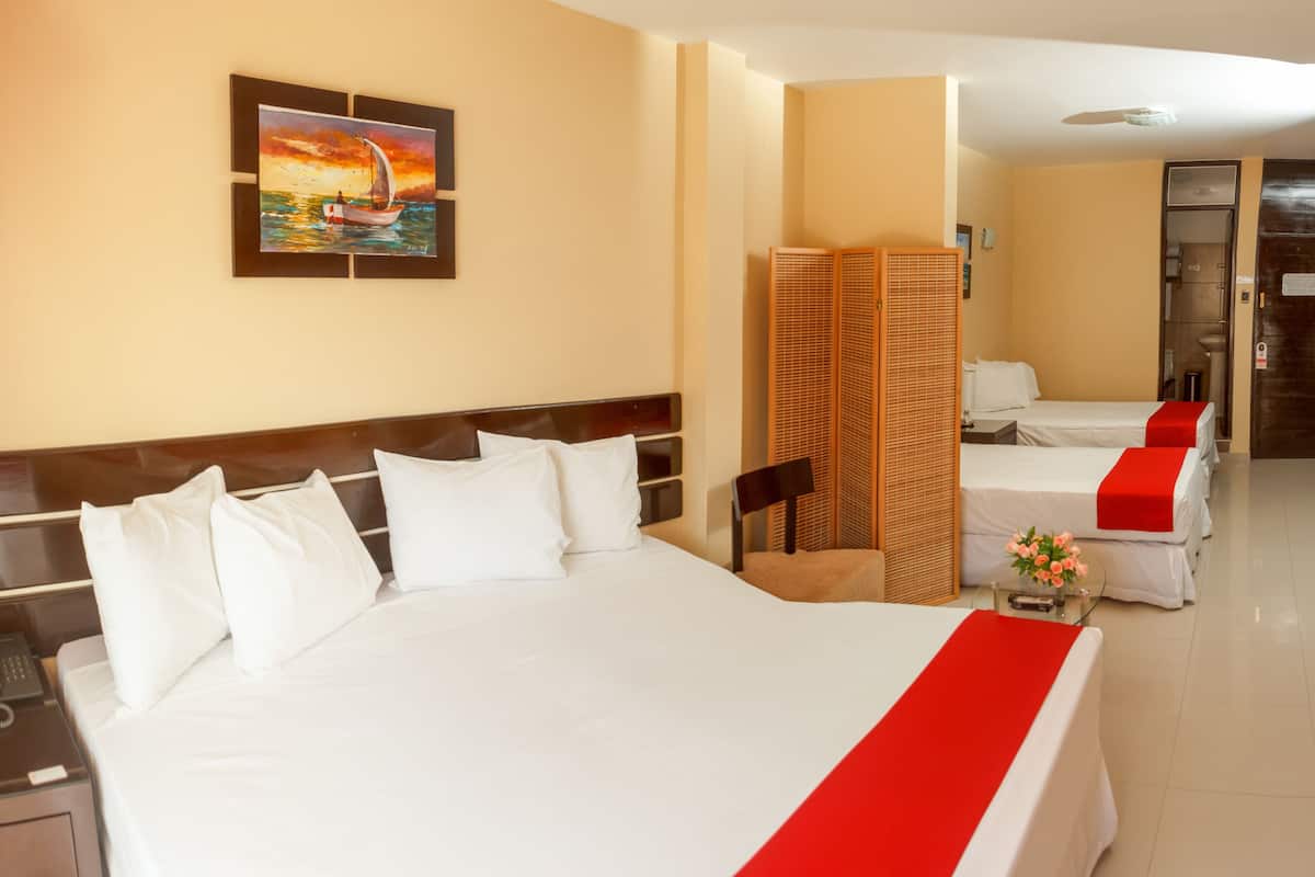 A hotel room with a large bed, a bedside table with a lamp, a phone, and a painting of a sunset over the sea. There is a red runner on the bed, and a small table with a vase of flowers.