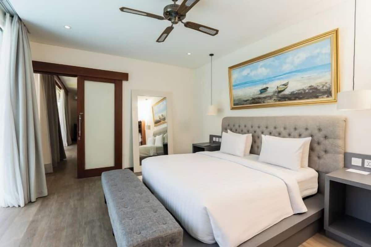 A hotel room with a bed, a bench, a ceiling fan, a painting of a beach, and a door leading to a bathroom.