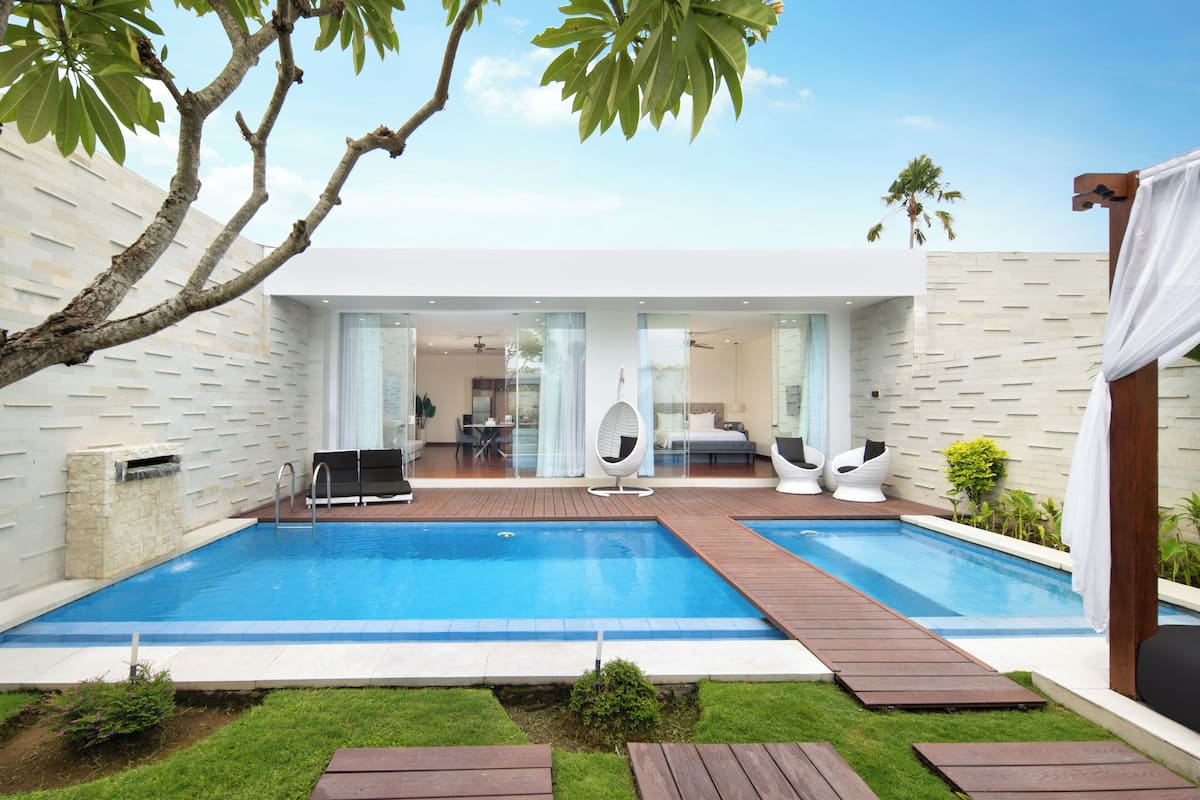 A modern outdoor pool area with a wooden deck, a glass-enclosed lounge, and a stone wall.