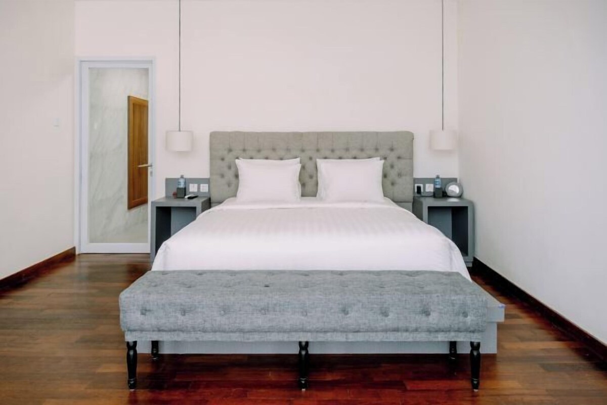 Un lit bien fait, agrémenté d’un banc recouvert d’un tissu gris, une table de chevet avec une lampe et une porte donnant sur une autre pièce.