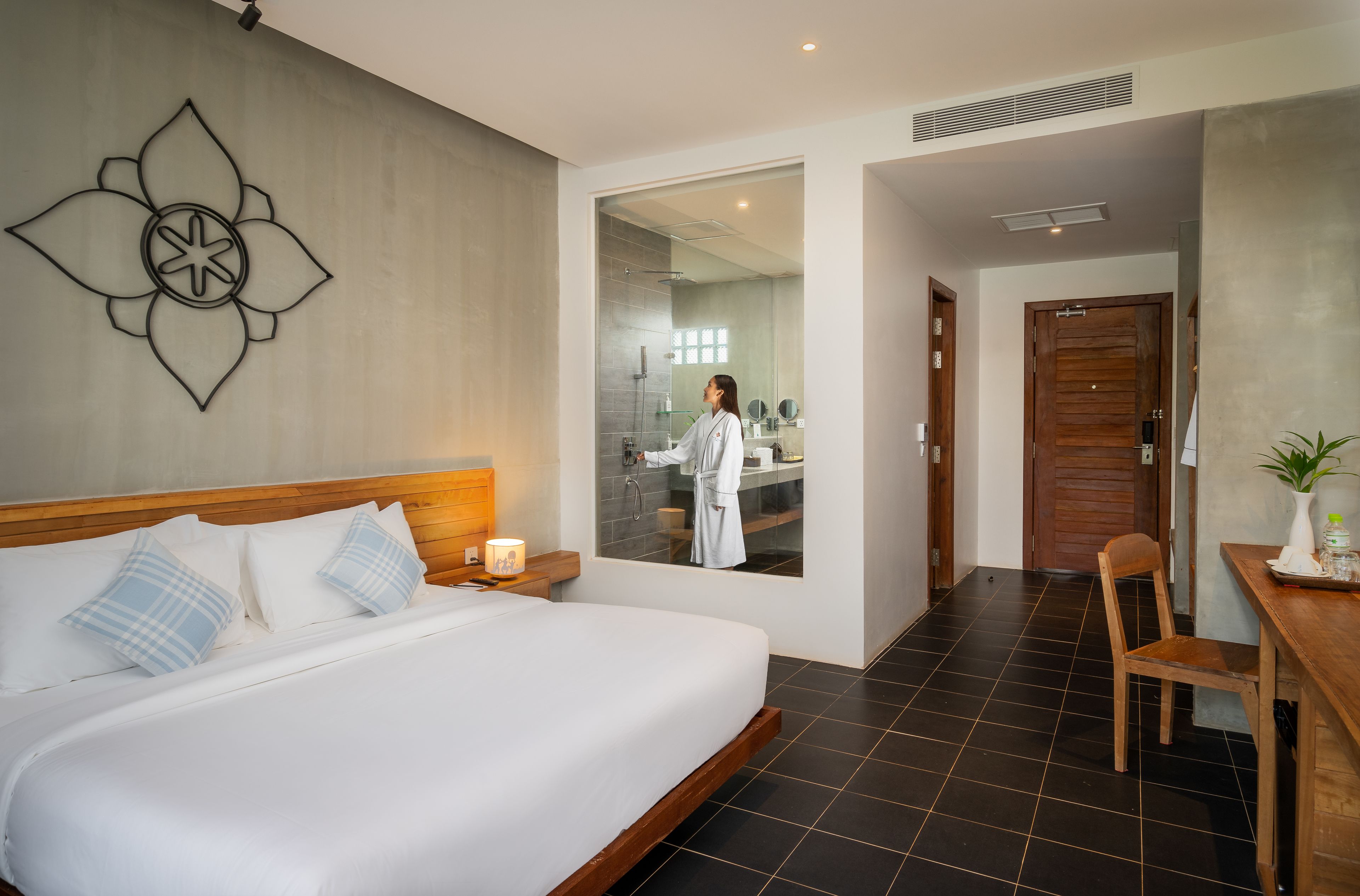 Ett modernt hotellrum med en stor säng, ett träfärgat sänggavel, en blomsterdekoration som hänger på väggen, ett badrum som syns genom en glasdörr och ett träbord med en stol.