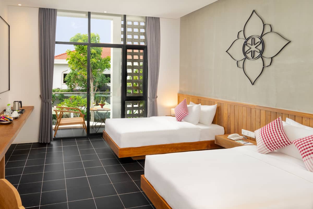 Habitación de hotel con dos camas, un escritorio, televisión y balcón con vistas a zonas verdes y edificios.
