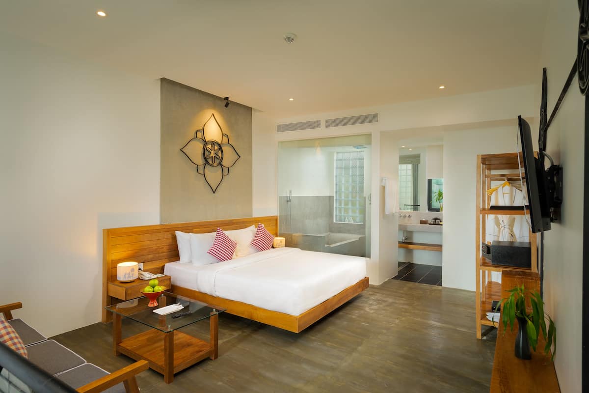 Habitación de hotel moderna con una cama de madera, una mesa de centro de vidrio, un televisor y un baño visible a través de una puerta de cristal.