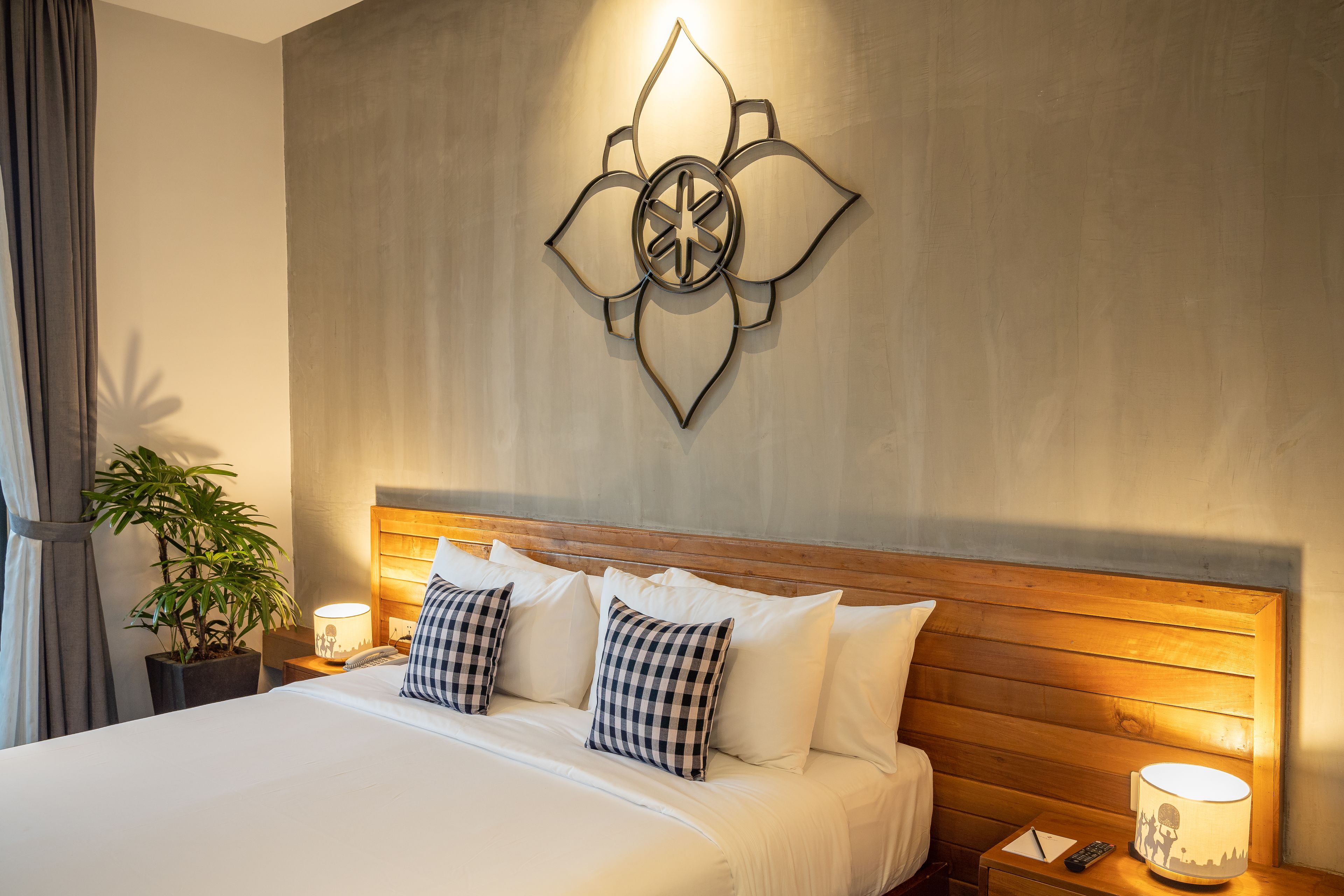 A neatly made bed with white linens and checkered pillows, a bedside table with a lamp, a wall-mounted decorative piece, and a potted plant.
