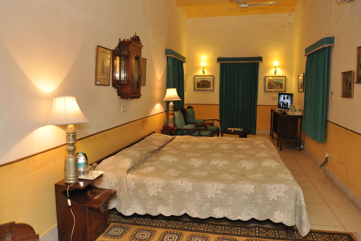 A hotel room with a bed, a desk with a lamp, a chair, a television, and a wall-mounted clock.