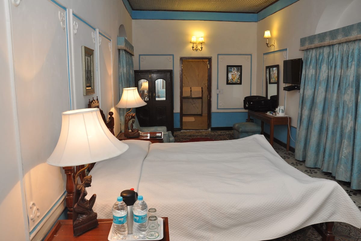 A hotel room with a bed, two bedside lamps, a nightstand with a lamp, a television, a desk with a chair, and a door leading to a bathroom.