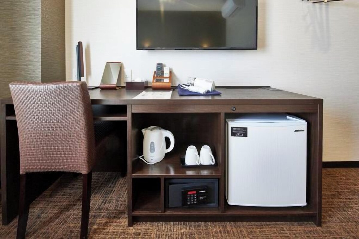 Une chambre d’hôtel équipée d’un bureau en bois, d’une télévision, d’un réfrigérateur, d’une bouilloire et d’une paire de chaussons.