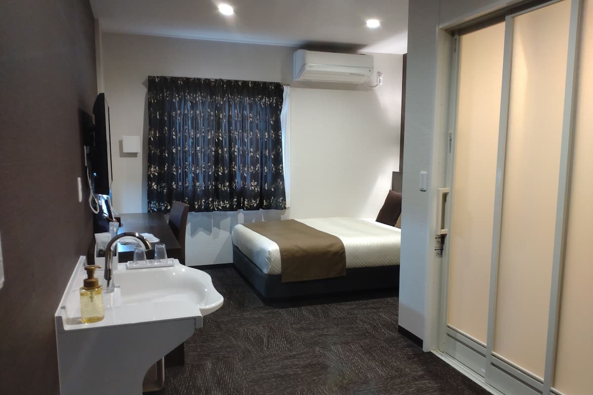 A compact hotel room with a single bed, a bedside table, a small desk, a sink with a mirror, and a wardrobe.