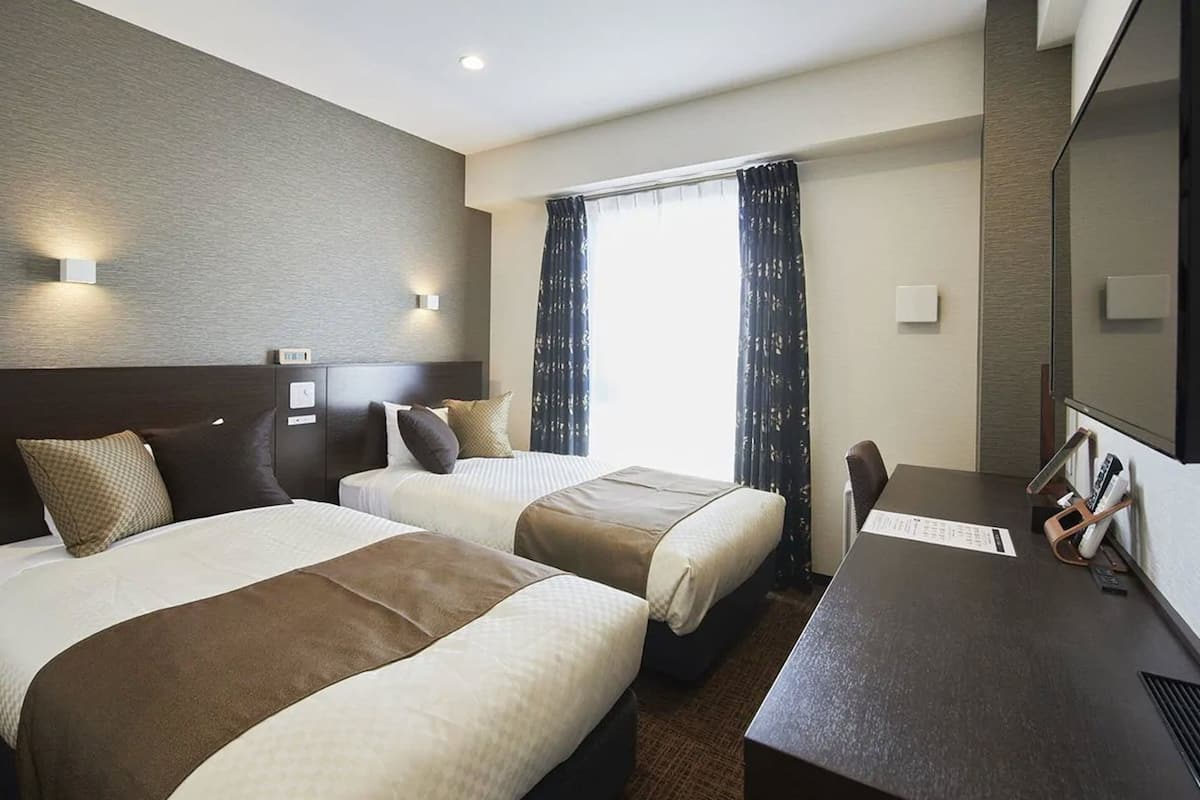 A hotel room with two beds, a desk, and a large window with curtains.