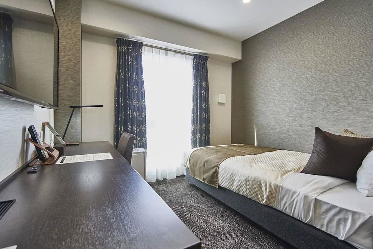 A hotel room with a bed, a desk with a chair, a TV, and a window with curtains.
