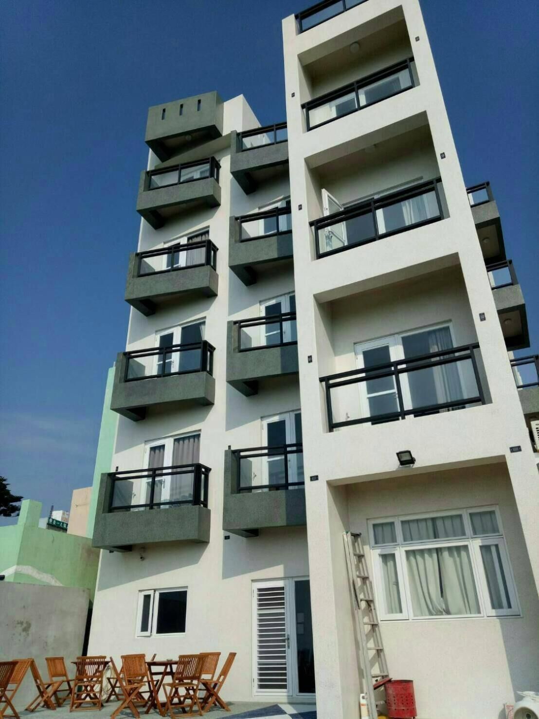 A multi-story residential building with balconies, a visible door, and a small outdoor seating area with chairs.