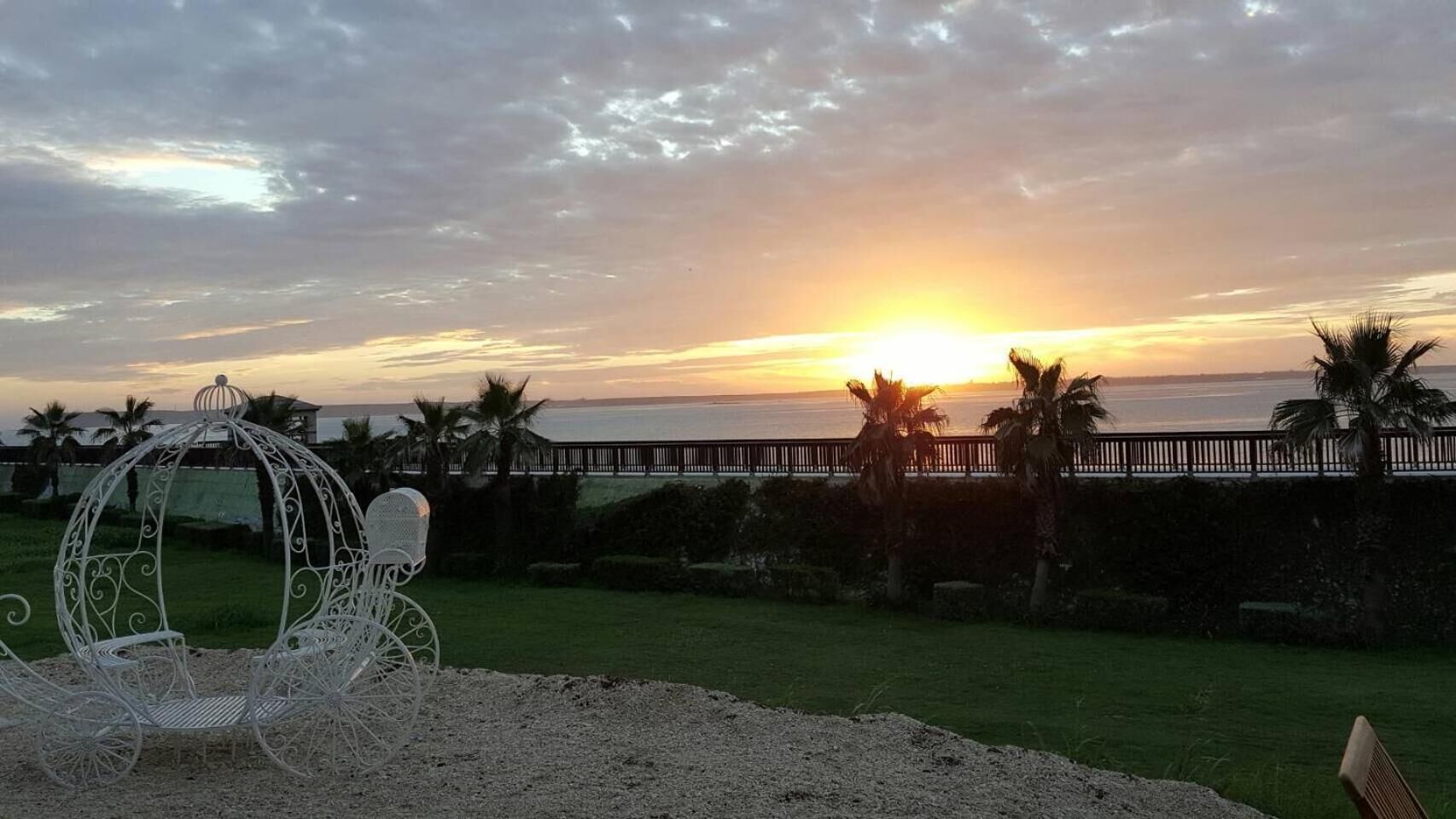 A sunset view with a white metal gazebo, palm trees, and a wooden fence overlooking a body of water.