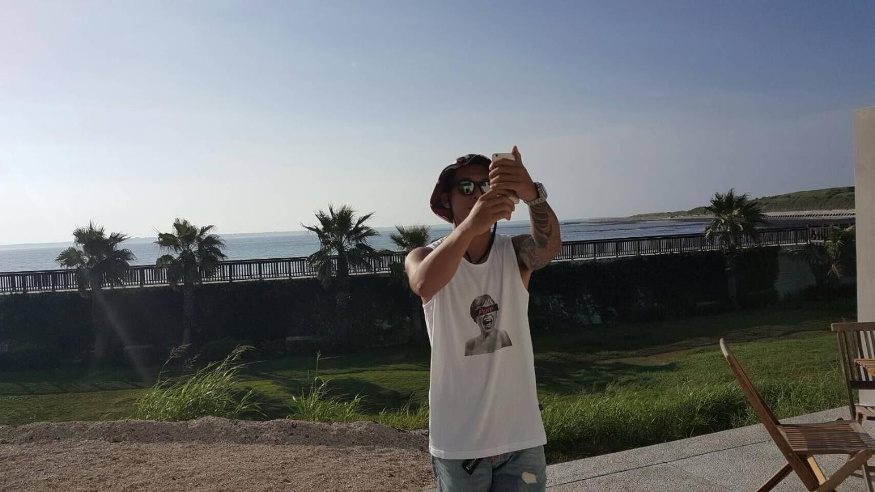 A person taking a photo with a smartphone, standing on a patio with a view of the ocean and palm trees.