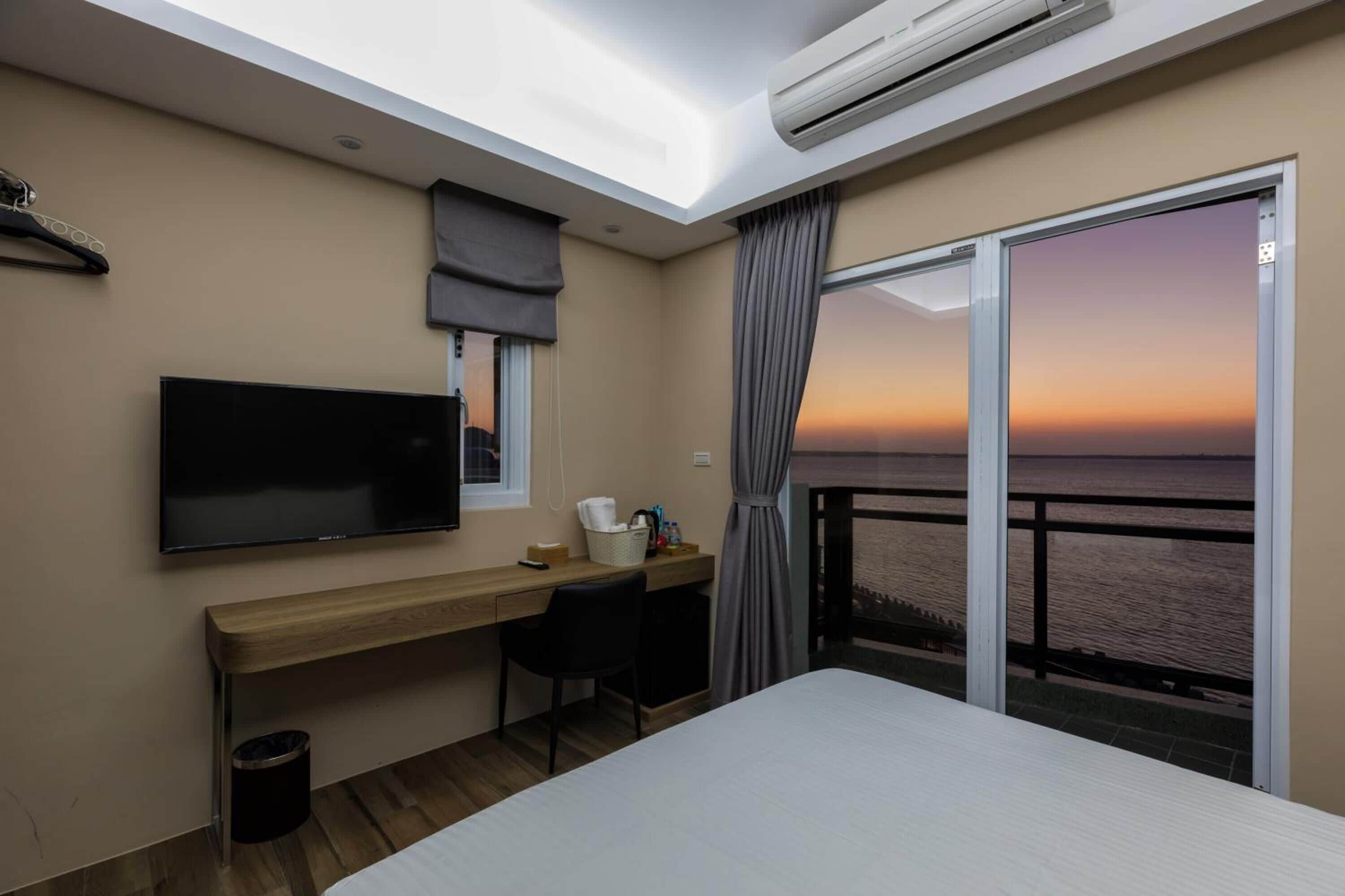 A hotel room with a bed, a desk, a television, and a large window with a view of the sea at sunset.