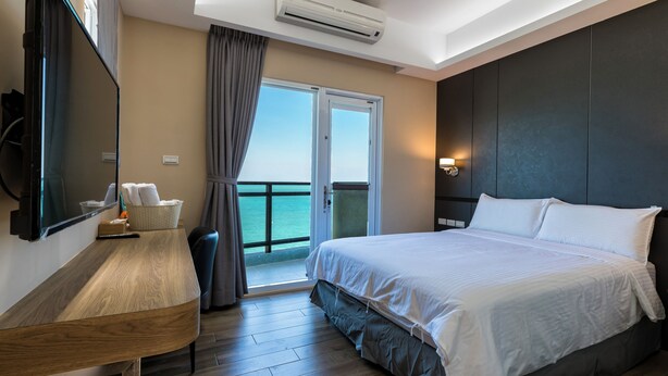 A hotel room with a bed, a desk, a chair, a television, an air conditioning unit, a window with curtains, and a view of the sea. - Jia Xiang Lin Guan Hai Homestay 2 (Magong)