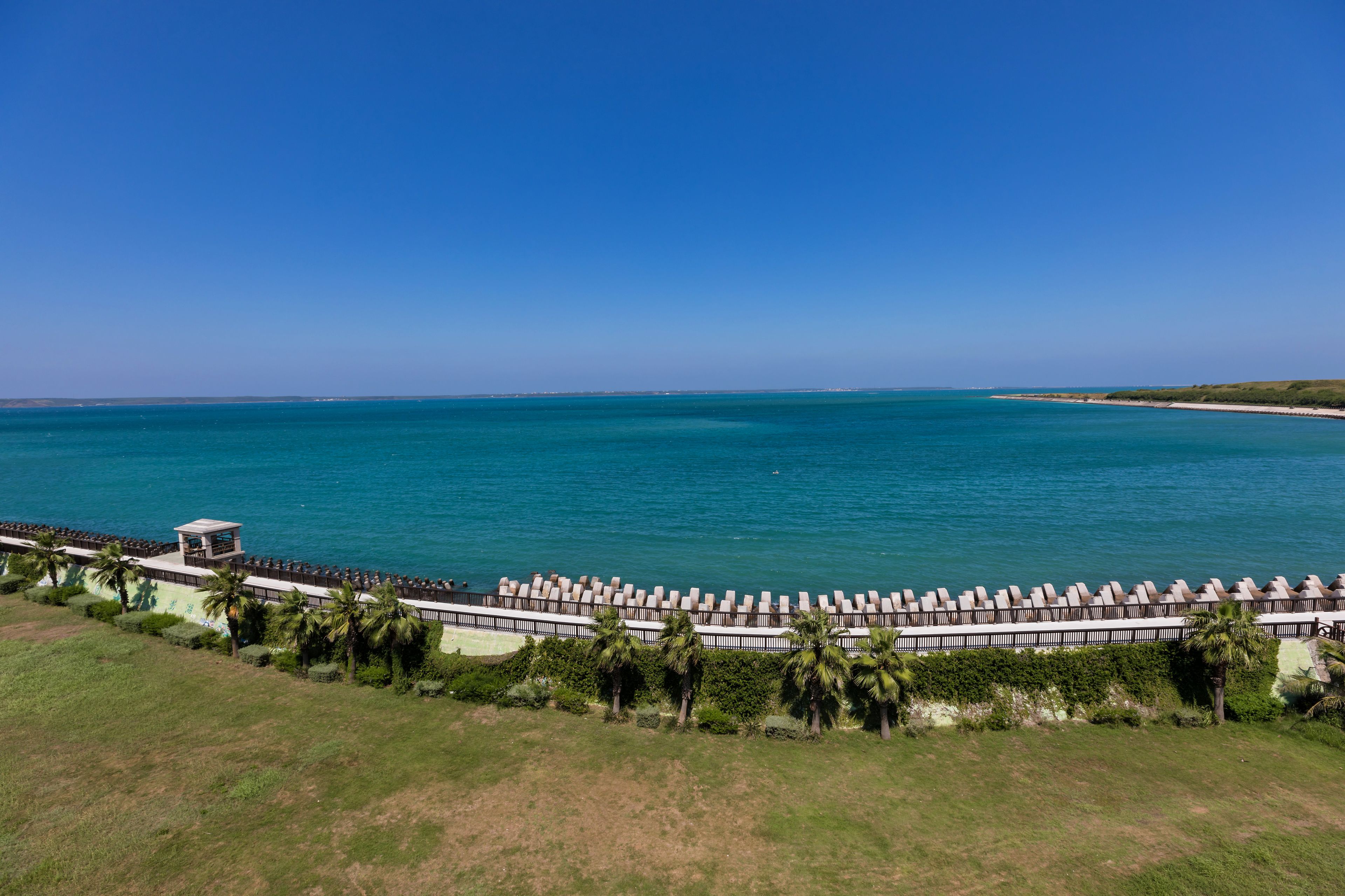 A coastal hotel with a long row of beachside rooms, a small tower structure, and a grassy area with palm trees.