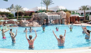 A swimming pool with people enjoying the water, a waterfall feature, and a landscaped area with palm trees and buildings in the background. - Verginia Sharm Resort & Aqua Park (Sharm El Sheikh)
