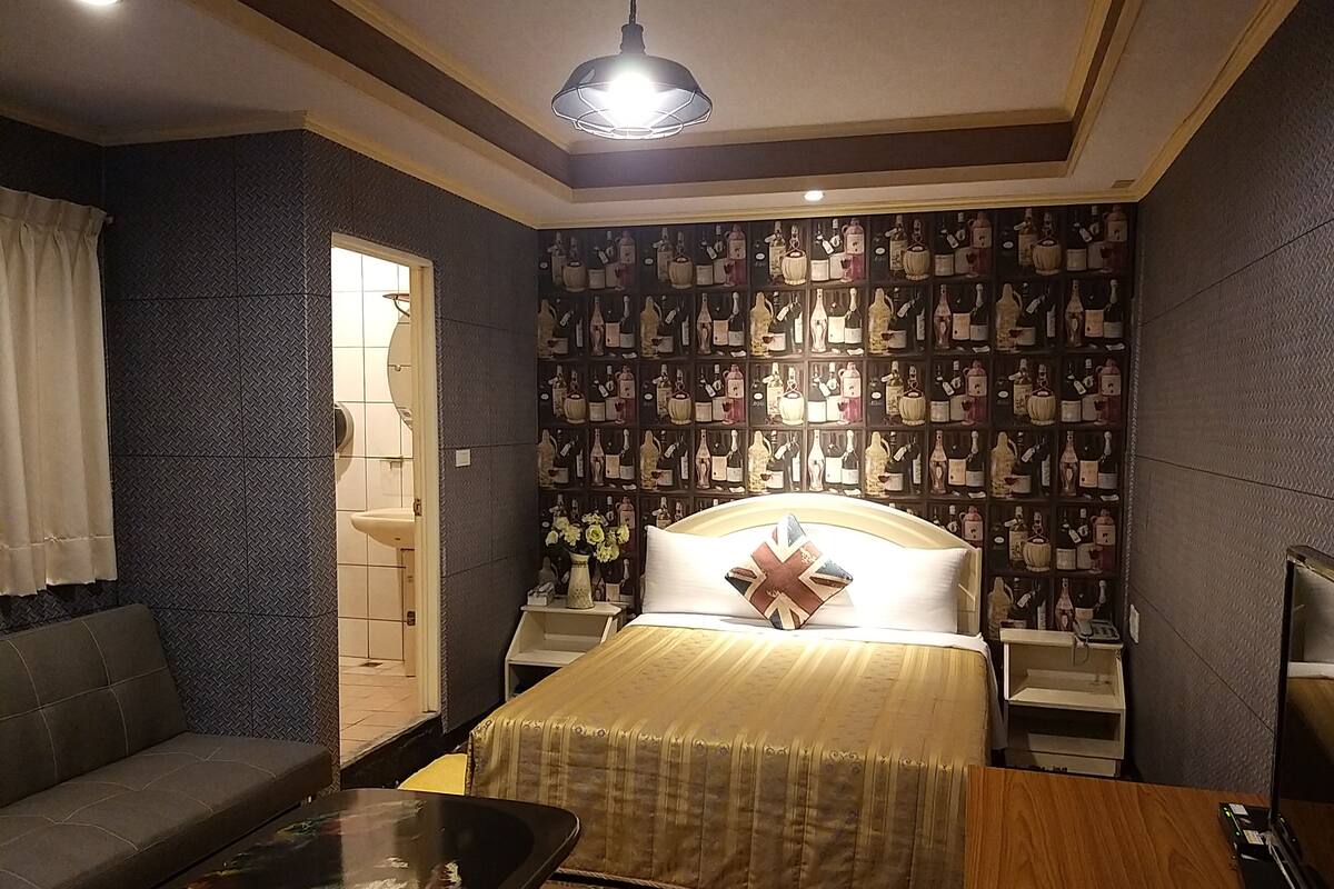 A hotel room with a bed, a sofa, a bedside table, a nightstand, and a wall with a pattern of bottles.