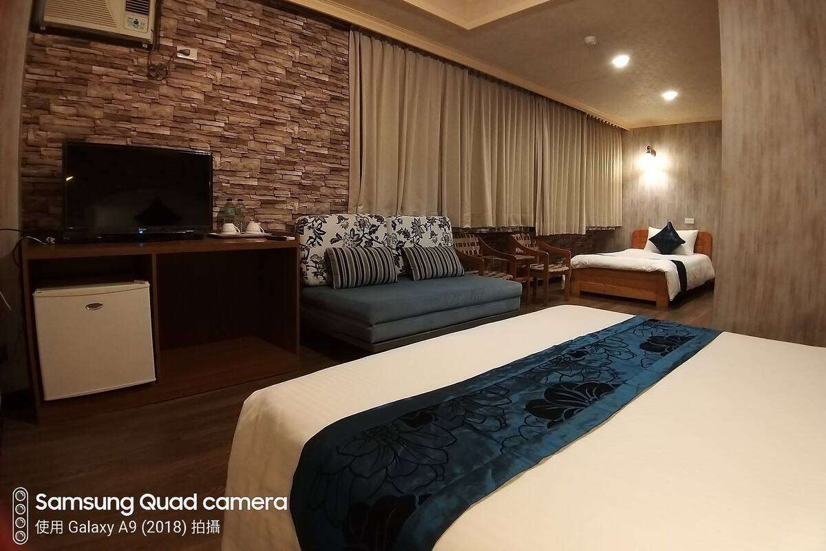 A hotel room with a stone wall, a flat-screen TV, a desk, a small fridge, a sofa, a bed, and a bedside table.