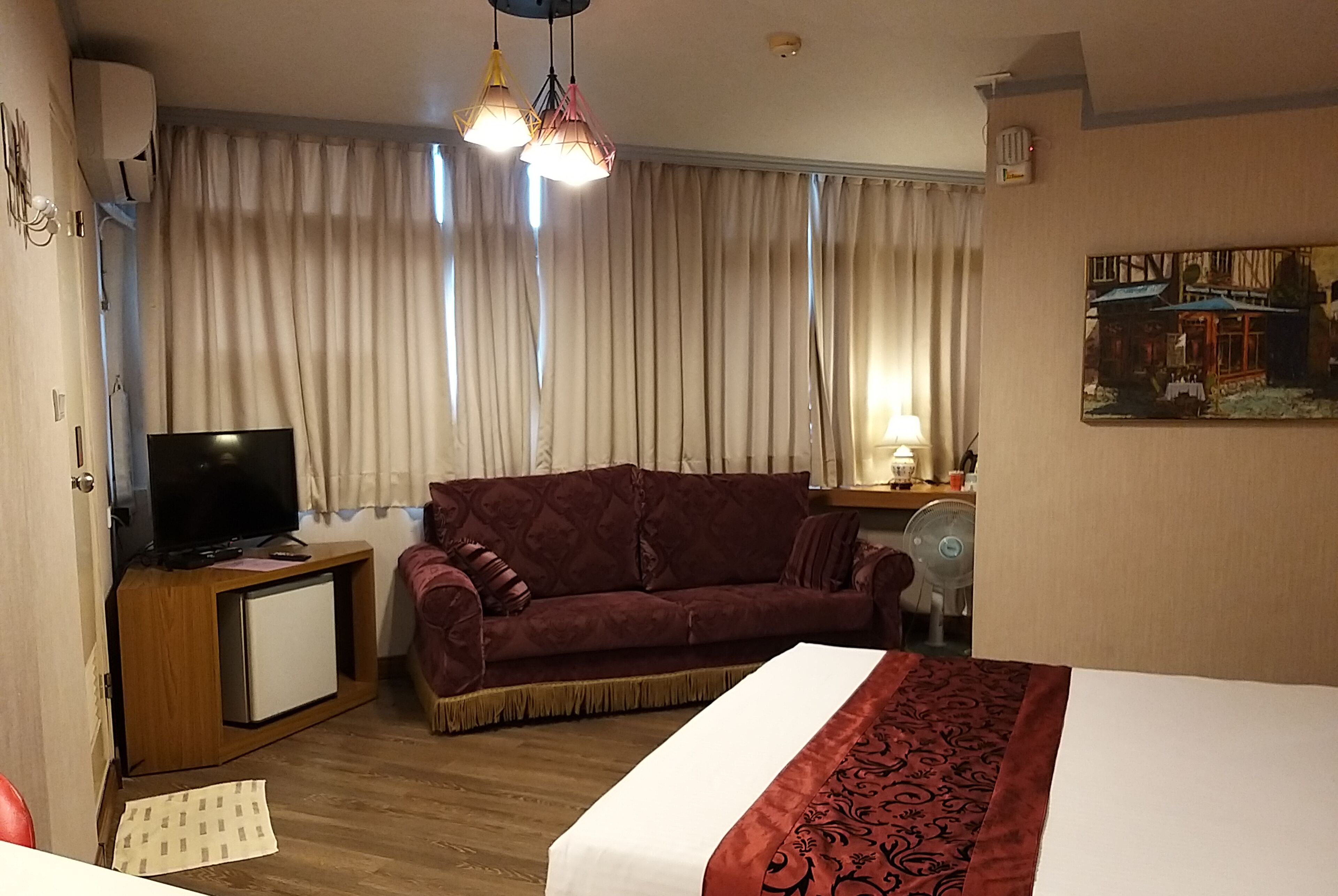 A hotel room with a bed, a TV on a wooden cabinet, a red sofa, a desk with a lamp, and a painting on the wall.