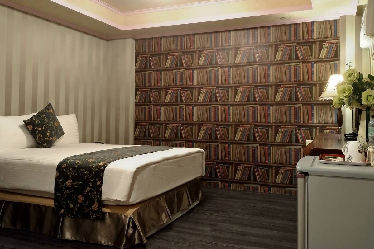 A hotel room with a large bookshelf, a bed with a floral pillow, and a desk with a vase of flowers.