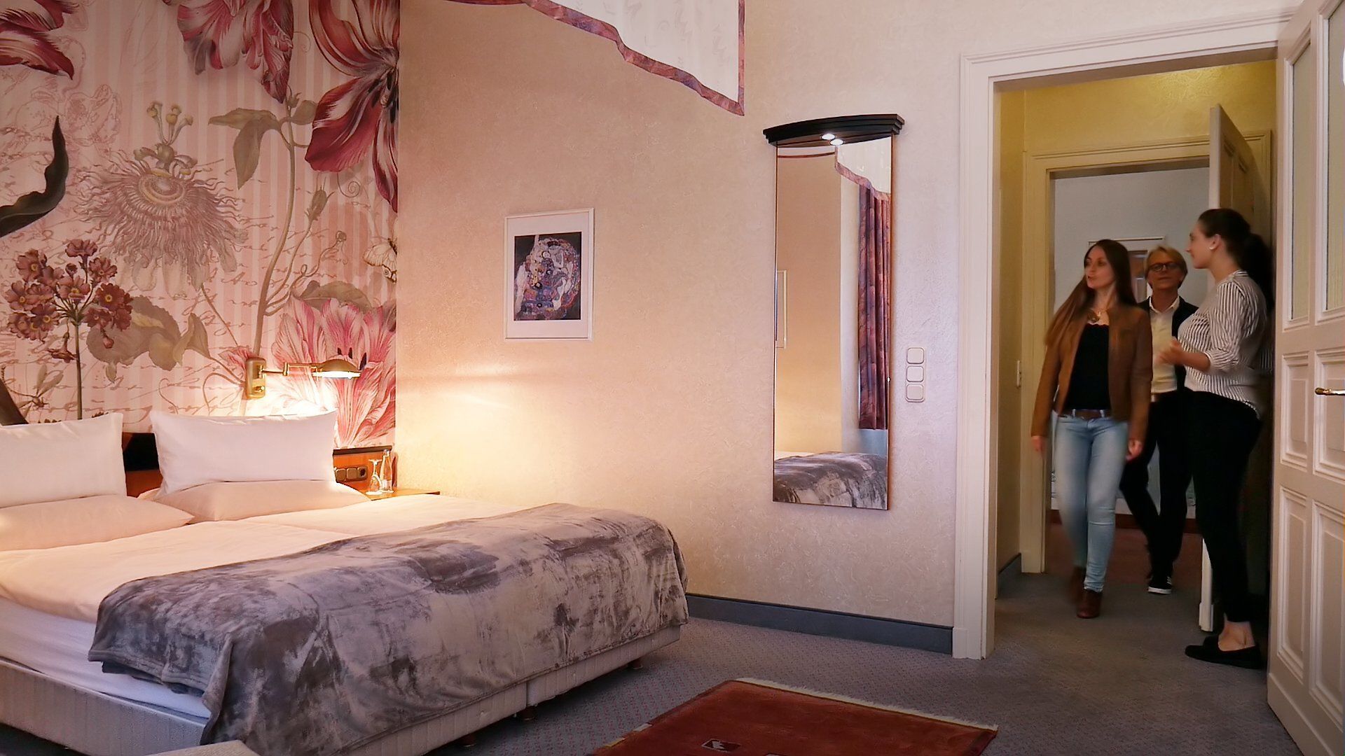A hotel room with a bed, a nightstand, a lamp, a mirror, and a door leading to another room.
