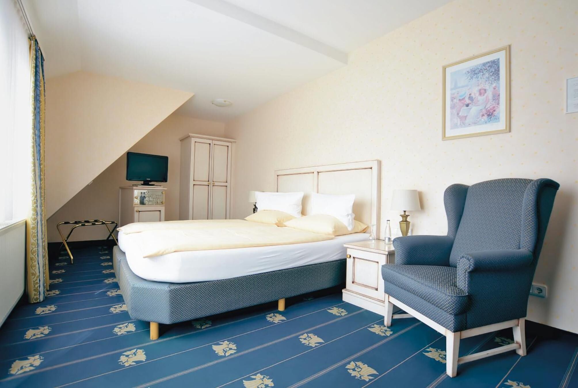 A hotel room with a bed, a chair, a nightstand, a television, and a painting on the wall.
