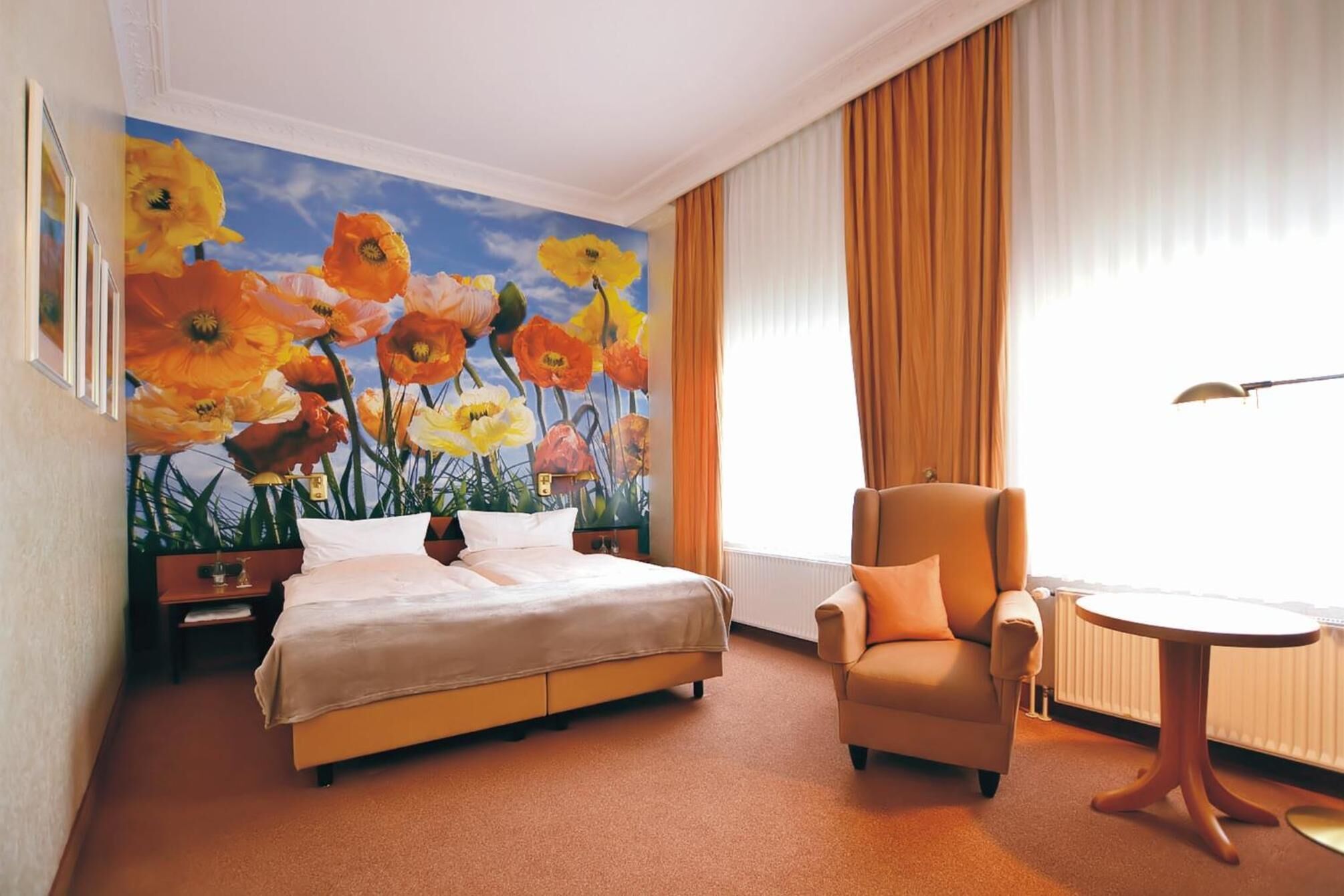 A hotel room with a bed, a chair, a small table, and a painting of flowers on the wall.