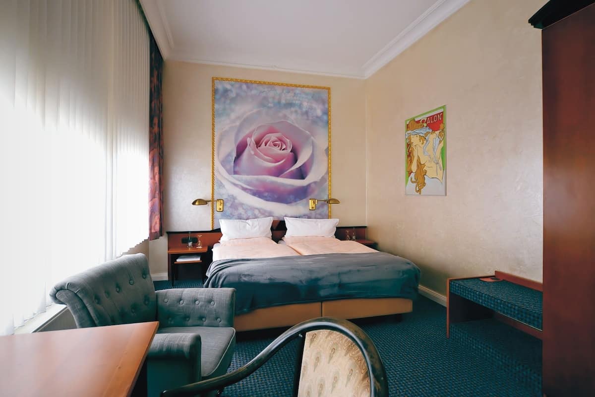 Ett hotellrum med en säng, ett skrivbord, en stol, ett fönster med persienner och en stor blomstermålning på väggen.