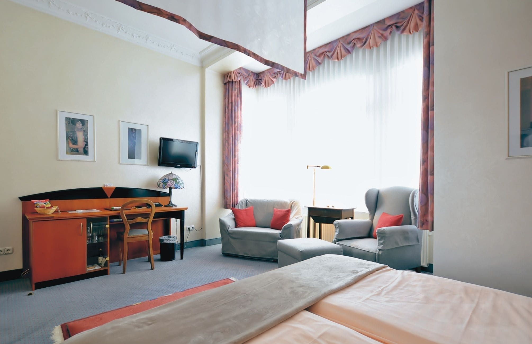 Hotellihuone, jossa on sänky, kaksi nojatuolia, työpöytä tietokoneella, televisio ja ikkuna, jossa on verhot.