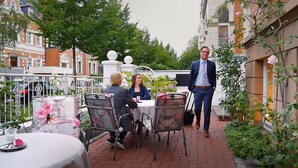 Un área de comedor al aire libre con mesas y sillas, rodeada de vegetación y edificios.