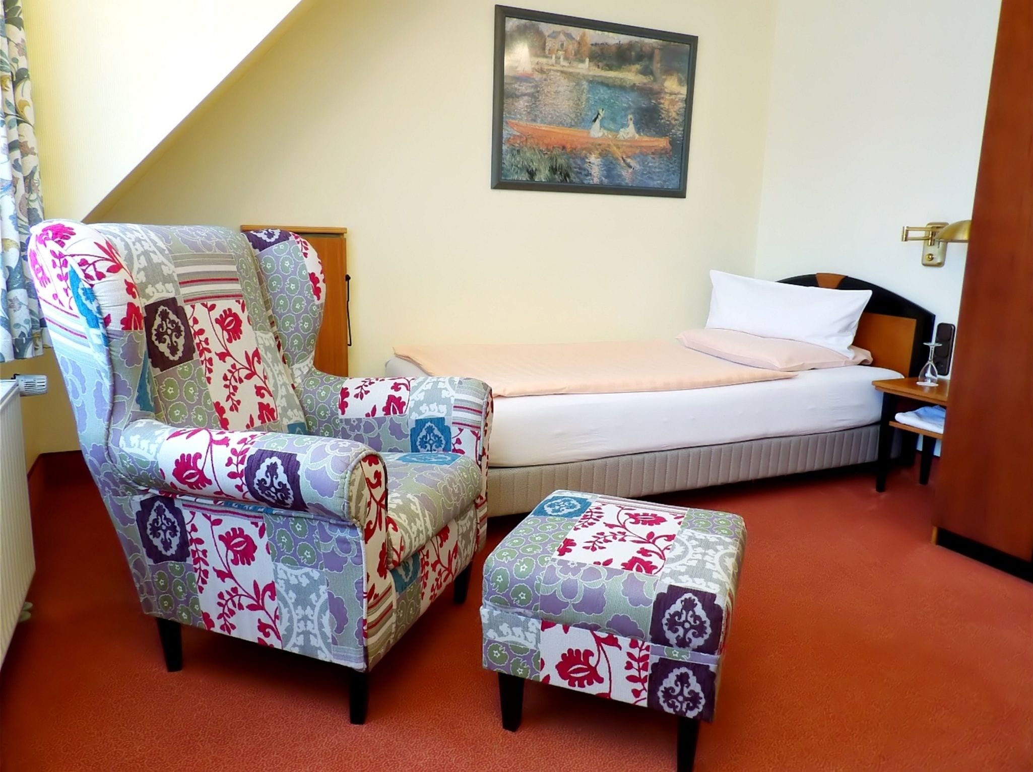 A room with a bed, a chair, an ottoman, a painting, and a radiator.