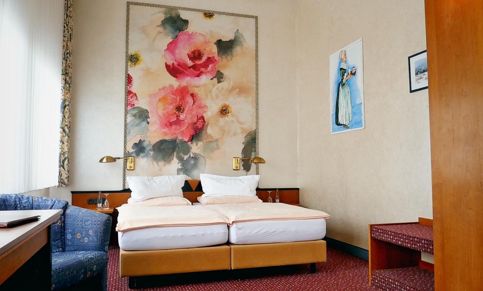 A hotel room with a bed, a desk, a chair, and a large floral painting on the wall.