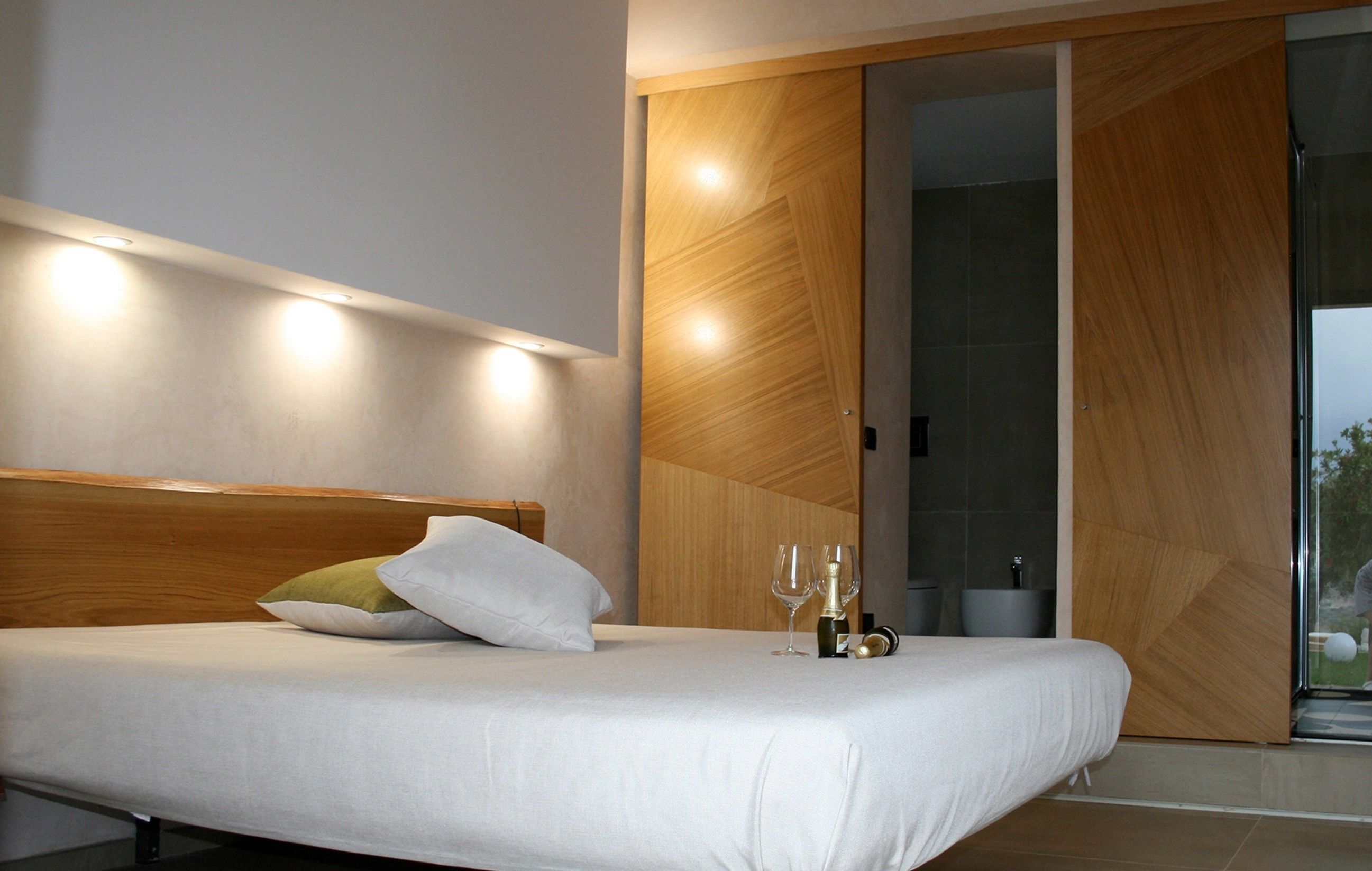 Una camera da letto moderna con una testiera in legno, un letto con lenzuola bianche, due cuscini e un comodino con due bicchieri da vino e una bottiglia.