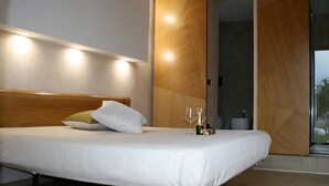 Un dormitorio moderno con cabecera de madera, cama con ropa blanca, dos almohadas y una mesita de noche con dos copas de vino y una botella.
