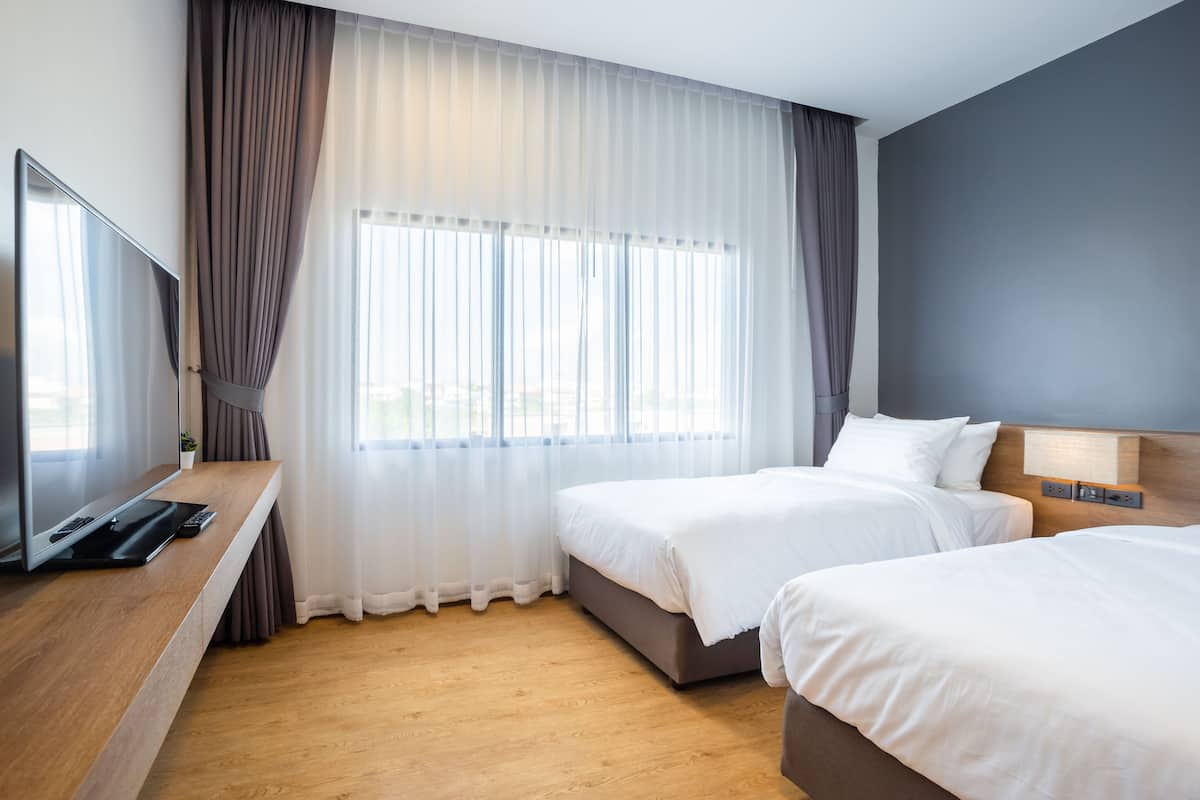 Een hotelkamer met twee bedden, een televisie op een houten bureau en een groot raam met dunne gordijnen.