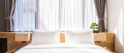 A hotel room with a large bed, two bedside tables, a nightstand, a lamp, and a view of the outside through sheer curtains.