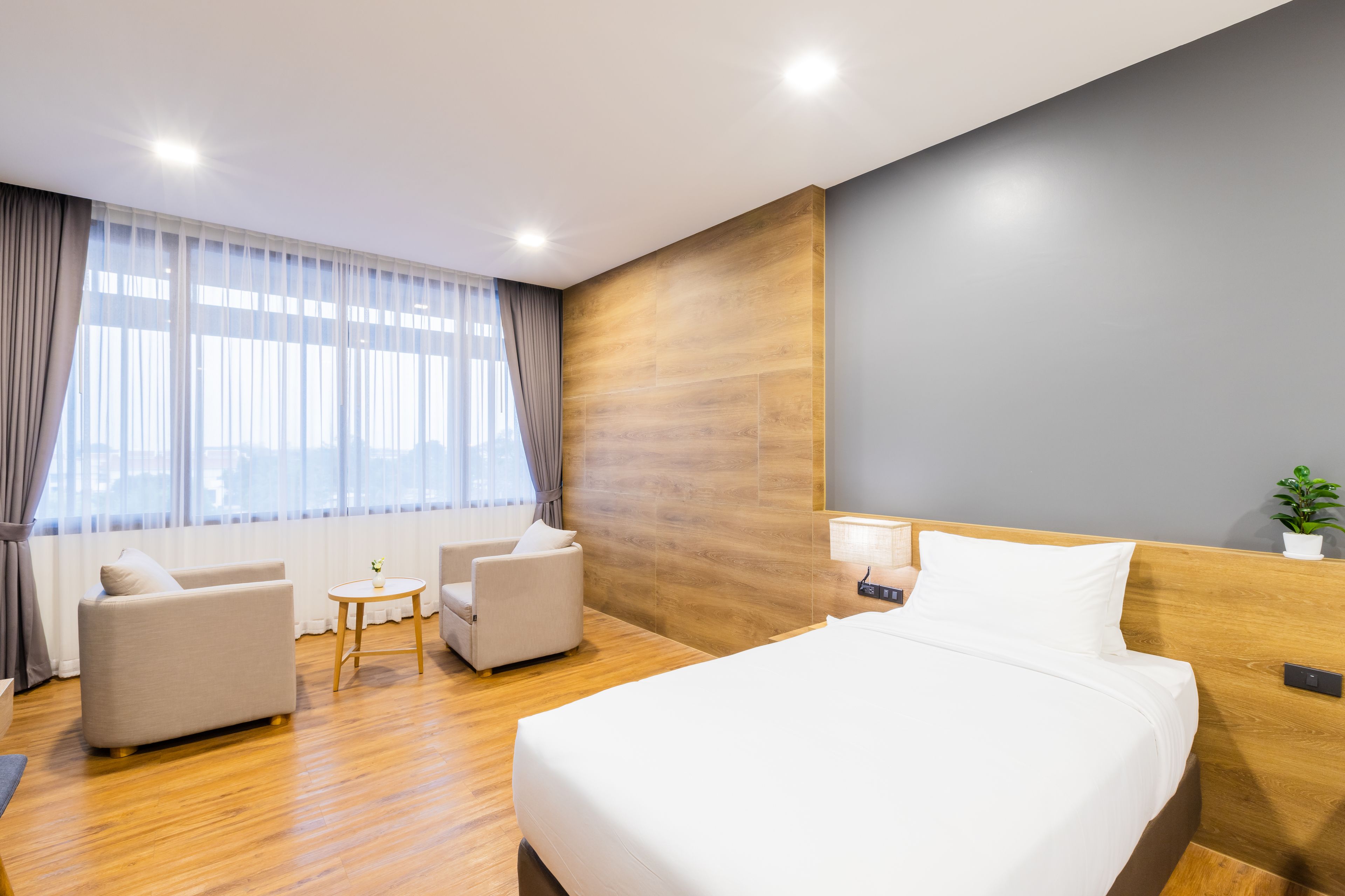 A modern hotel room with a large bed, two armchairs, a small table, and a large window with curtains.