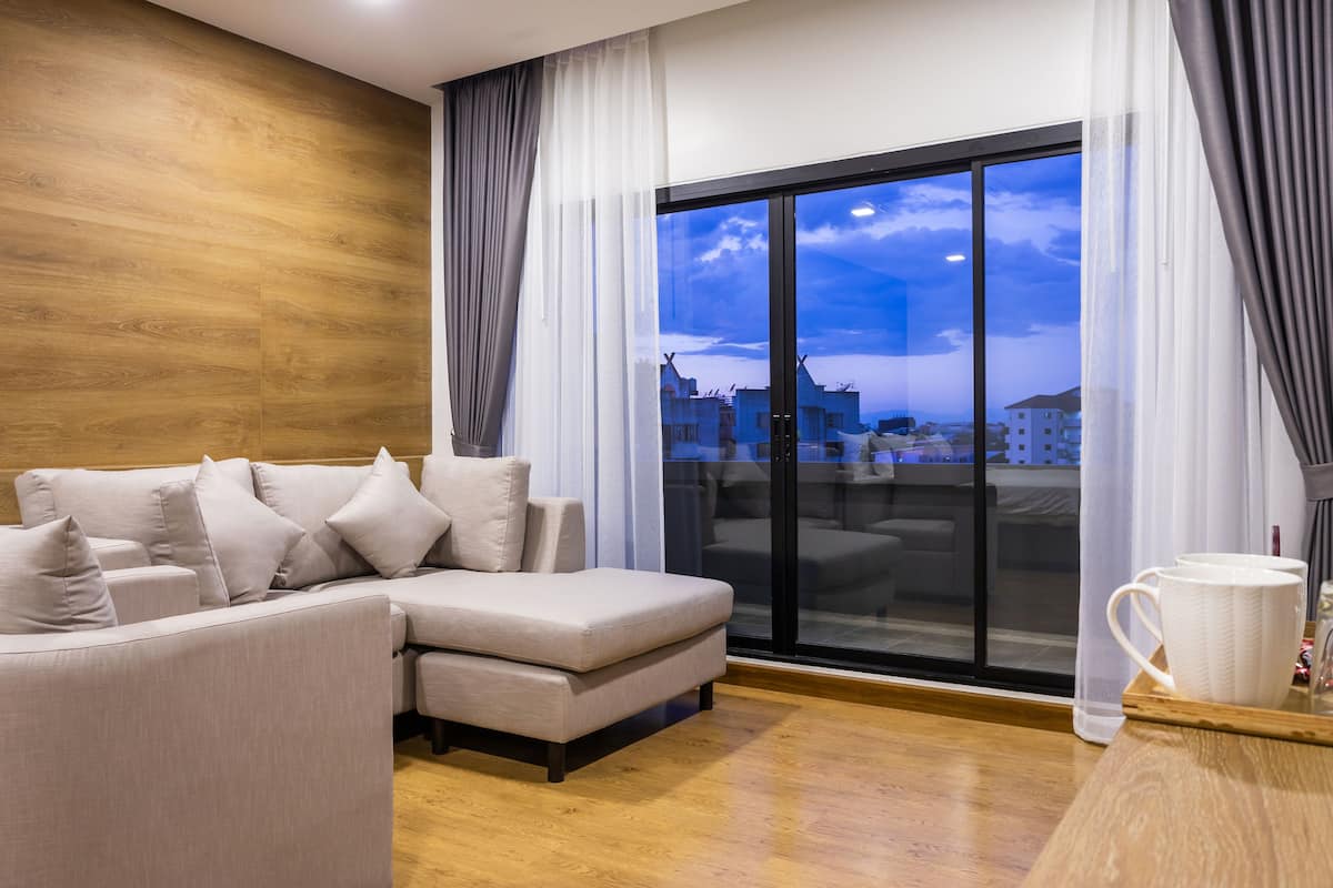 Een moderne woonkamer met een bank, een groot raam met gordijnen en uitzicht op gebouwen.