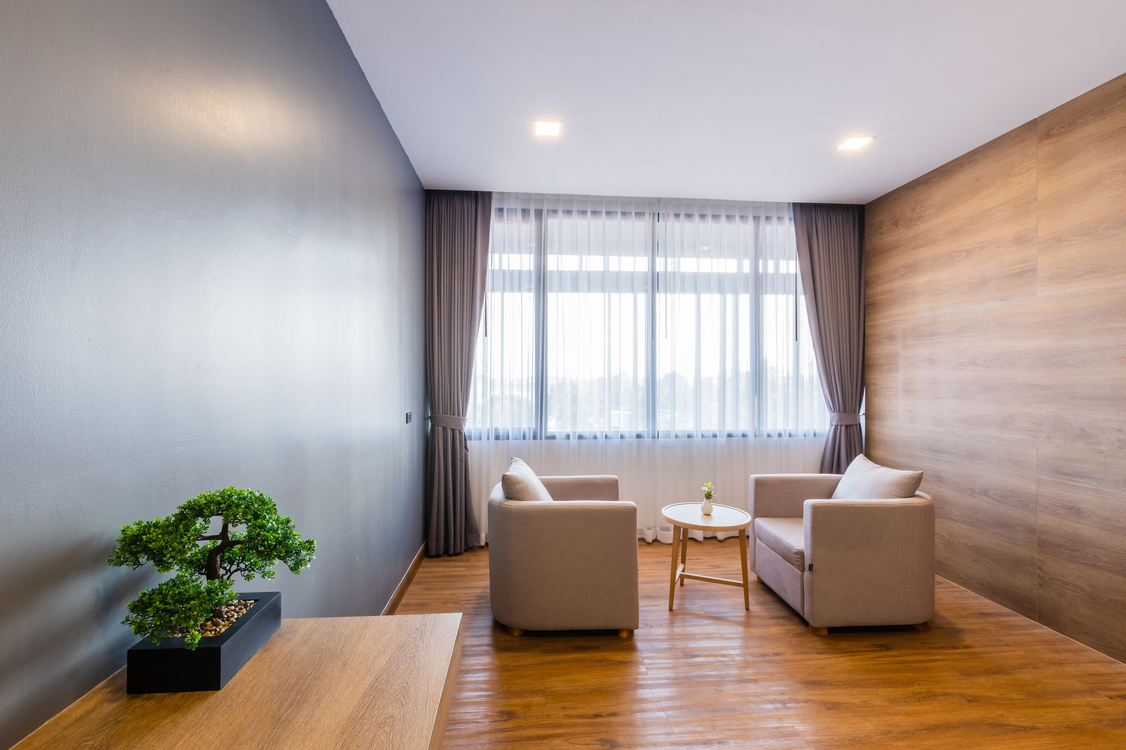 A modern room with a wooden floor, two beige armchairs, a small round table, and a bonsai tree in a black planter.